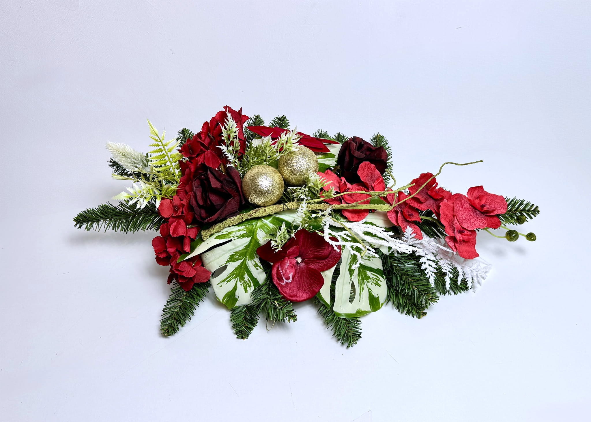 Podłużny stroik bożonarodzeniowy w tradycyjnej kolorystyce czerwieni, zieleni i złota. Baza to gęste, sztuczne gałązki świerkowe. Dekorację tworzą intensywnie czerwone róże i inne kwiaty, a w centrum widoczne są dwie złote, brokatowe bombki. Kompozycja zawiera duże liście w odcieniach głębokiej zieleni z białym wzorem (warygacją), przeplatane oszronionymi białymi gałązkami.