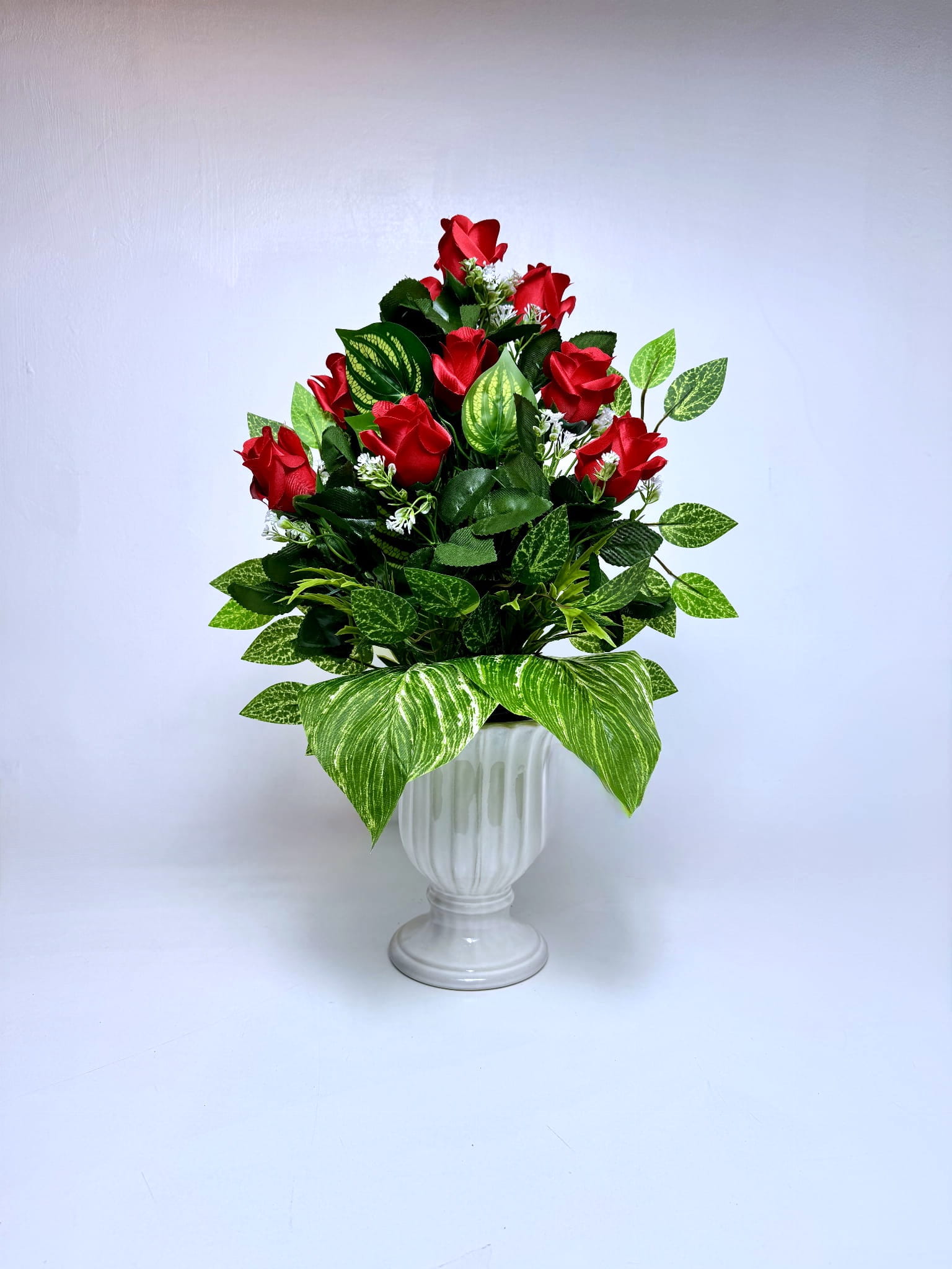 Zdjęcie przedstawia wysoki, stożkowaty bukiet nagrobny umieszczony w jasnoszarym, dekoracyjnym wazonie. Kompozycja składa się z wielu pąków sztucznych róż w kolorze głębokiej czerwieni, które są rozmieszczone kaskadowo – od pojedynczego kwiatu na szczycie po szerszą podstawę. Bukiet jest gęsto wypełniony zielonymi liśćmi o różnych kształtach, w tym dużymi, pasiastymi liśćmi u dołu oraz mniejszymi gałązkami z białymi kwiatuszkami gipsówki.