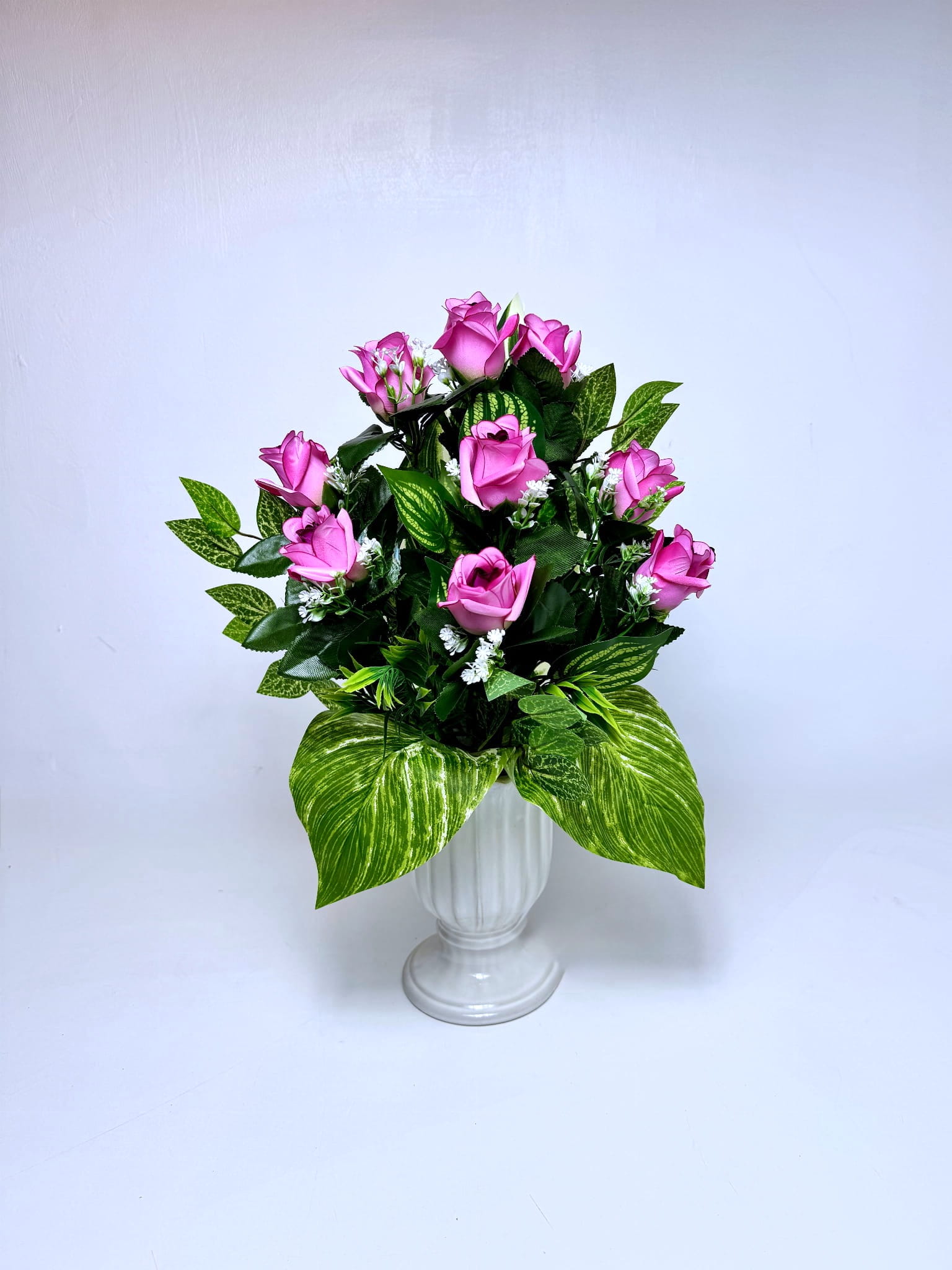 Kompozycja do wazonu składająca się z różowych pąków róż rozmieszczonych na różnych wysokościach. Kwiaty otoczone są gęstymi, zielonymi liśćmi – mniejszymi w górnej części i dużymi, pasiastymi liśćmi u podstawy. Pomiędzy różami widoczne są drobne białe akcenty kwiatowe. Bukiet ma kształt stożka i jest osadzony w czarnym, stabilnym wkładzie wazonowym. Całość prezentuje się naturalnie i świeżo. Wymiary dekoracji to 50 cm wysokości oraz 35 cm szerokości.