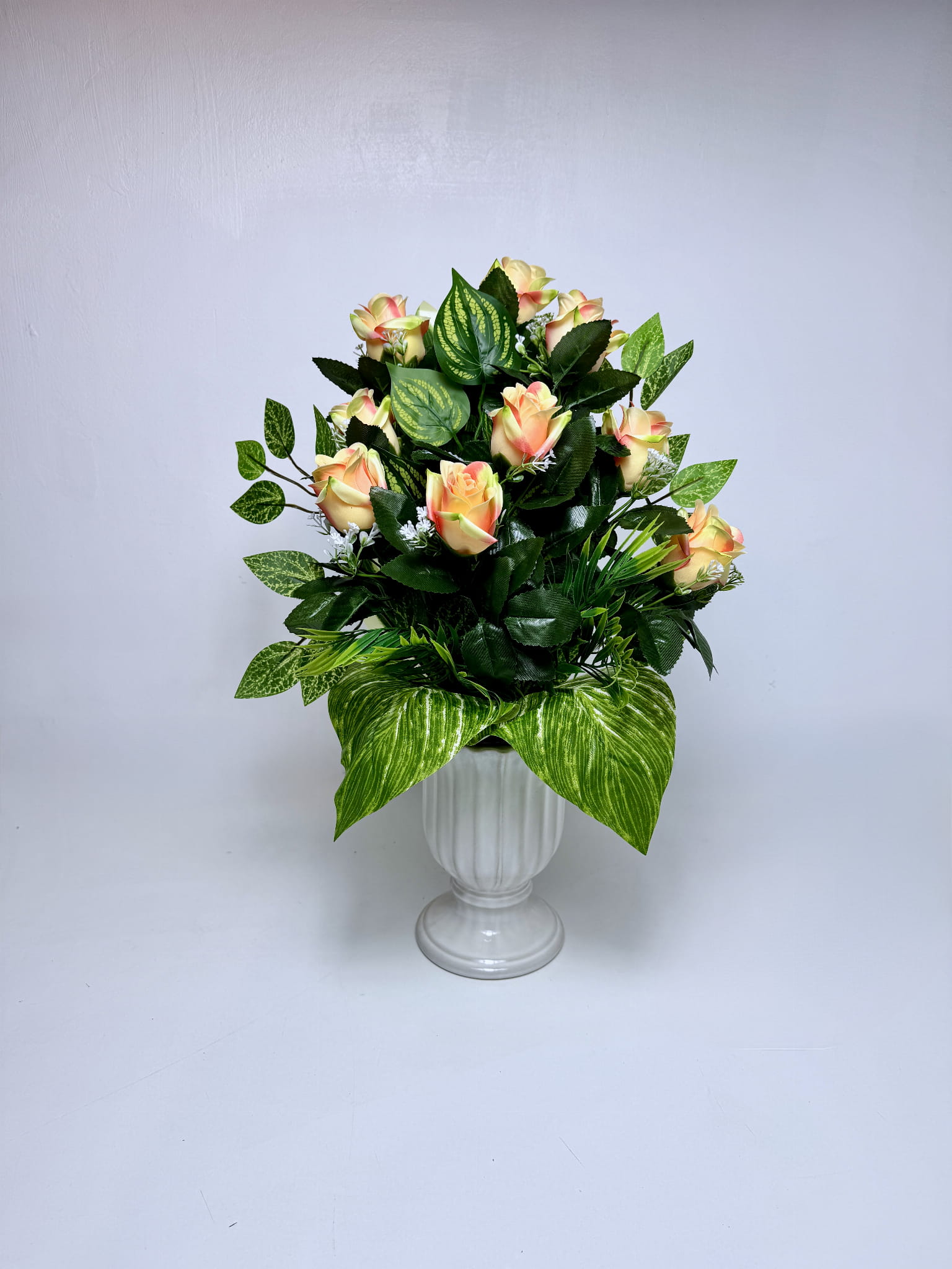 Kompozycja do wazonu w kształcie smukłego stożka, z pąkami róż w kolorze kremowo-brzoskwiniowym z różowymi krawędziami. Kwiaty są gęsto rozmieszczone wśród ciemnozielonych liści róż i jaśniejszych, wzorzystych liści dekoracyjnych. U podstawy bukietu znajdują się duże, szerokie liście o sercowatym kształcie z wyraźnymi jasnymi pasami. Między różami widoczne są drobne białe kwiatuszki. Całość osadzona jest w czarnym wkładzie wazonowym. Wymiary: 50 cm wysokości, 35 cm szerokości.