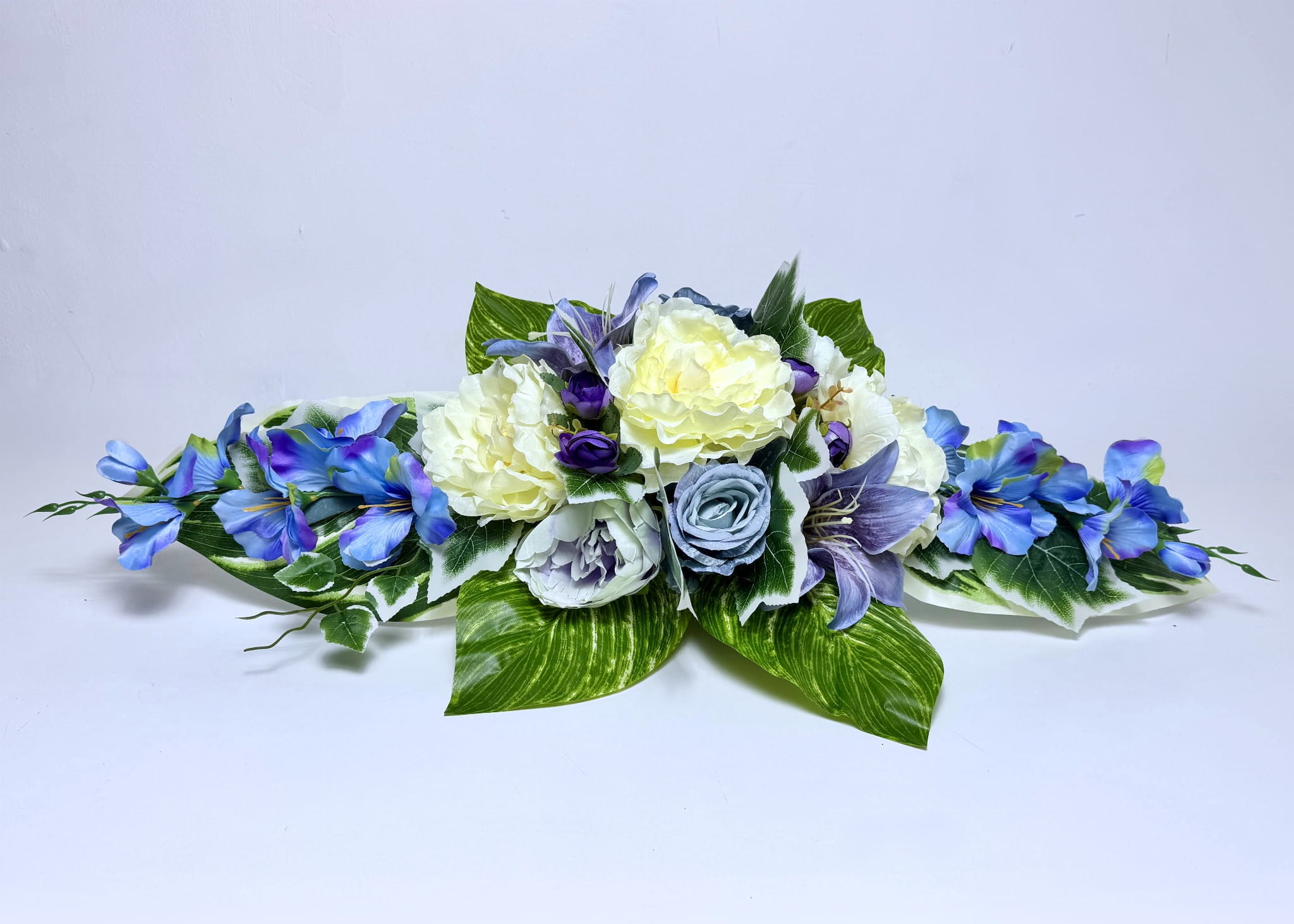 Podłużna, pozioma kompozycja nagrobna o długości 80 cm. W centrum znajdują się duże, kremowe peonie oraz jedna szaroniebieska róża. Po bokach umieszczono błękitne lilie o wywiniętych płatkach oraz małe, fioletowe akcenty. Całość wypełniona jest różnorodnymi liśćmi: dużymi z białym wzorem oraz drobnymi gałązkami bluszczu. Stroik ma niską, rozłożystą formę. Podstawa jest dociążona masą betonową, co zapewnia stabilność na pomniku. Kolorystyka łączy biel, błękit, fiolet i zieleń.