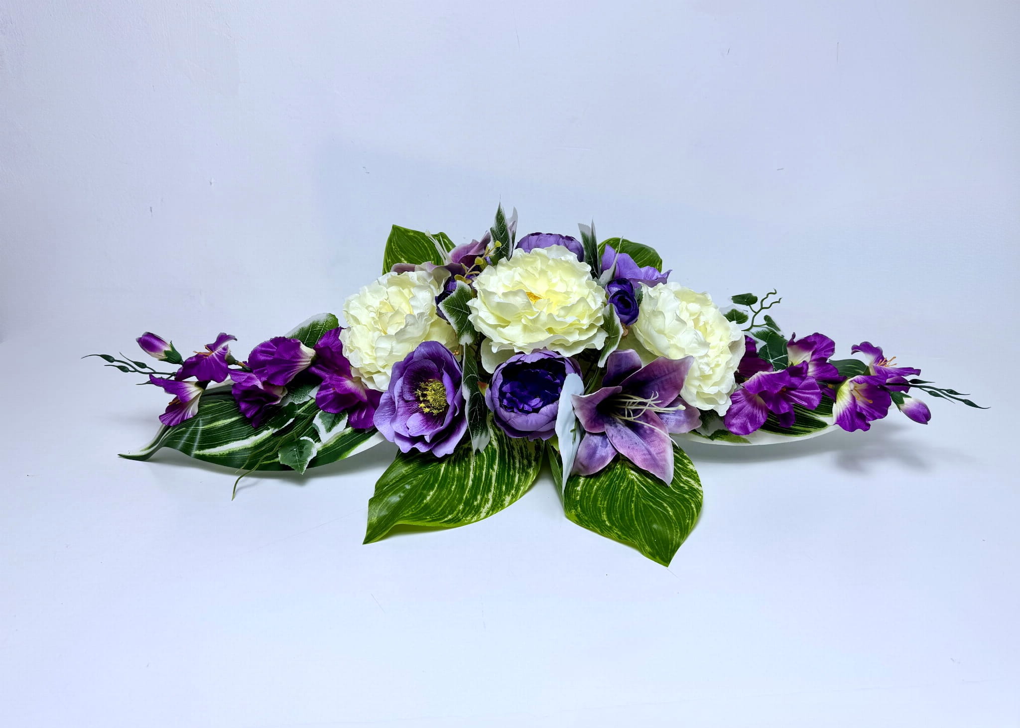 Pozioma, dociążona kompozycja nagrobna o długości 80 cm. W centrum znajdują się trzy duże kremowe peonie otoczone fioletowymi kwiatami o ciemnych środkach oraz fioletową lilią. Boki stroika zdobią rozłożyste, fioletowe gałązki kwiatowe. Całość spoczywa na bazie z dużych, zielonych liści z jasnym, pasiastym wzorem oraz mniejszych listków bluszczu. Podstawa wypełniona jest masą betonową, co zapewnia stabilność. Kolorystyka łączy jasny krem, intensywny fiolet i soczystą zieleń.