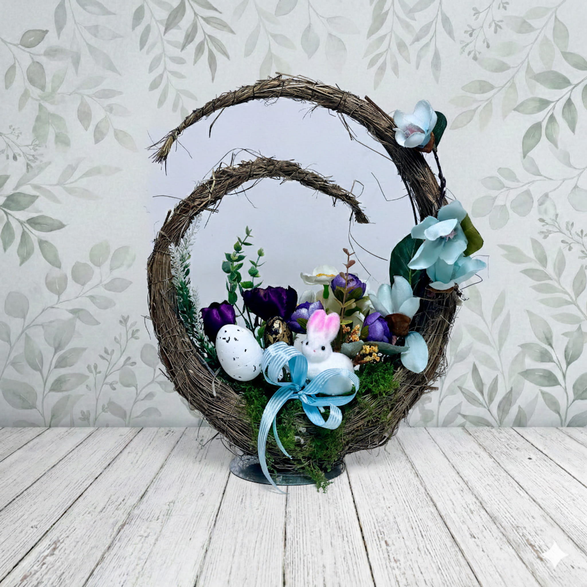 Mały, biały koszyczek z uchwytem wypełniony wiosennymi kwiatami w odcieniach błękitu, bieli i jasnej żółci. Wewnątrz znajdują się pąki tulipanów oraz drobne bratki, otoczone gęstymi, zielonymi liśćmi i listkami paproci. Koszyk ma strukturę plecionki i jest dociążony masą betonową, co zapewnia mu stabilność na pomniku. Kompozycja jest zwarta i lekka wizualnie. Wymiary dekoracji to 33 cm wysokości oraz 26 cm szerokości.