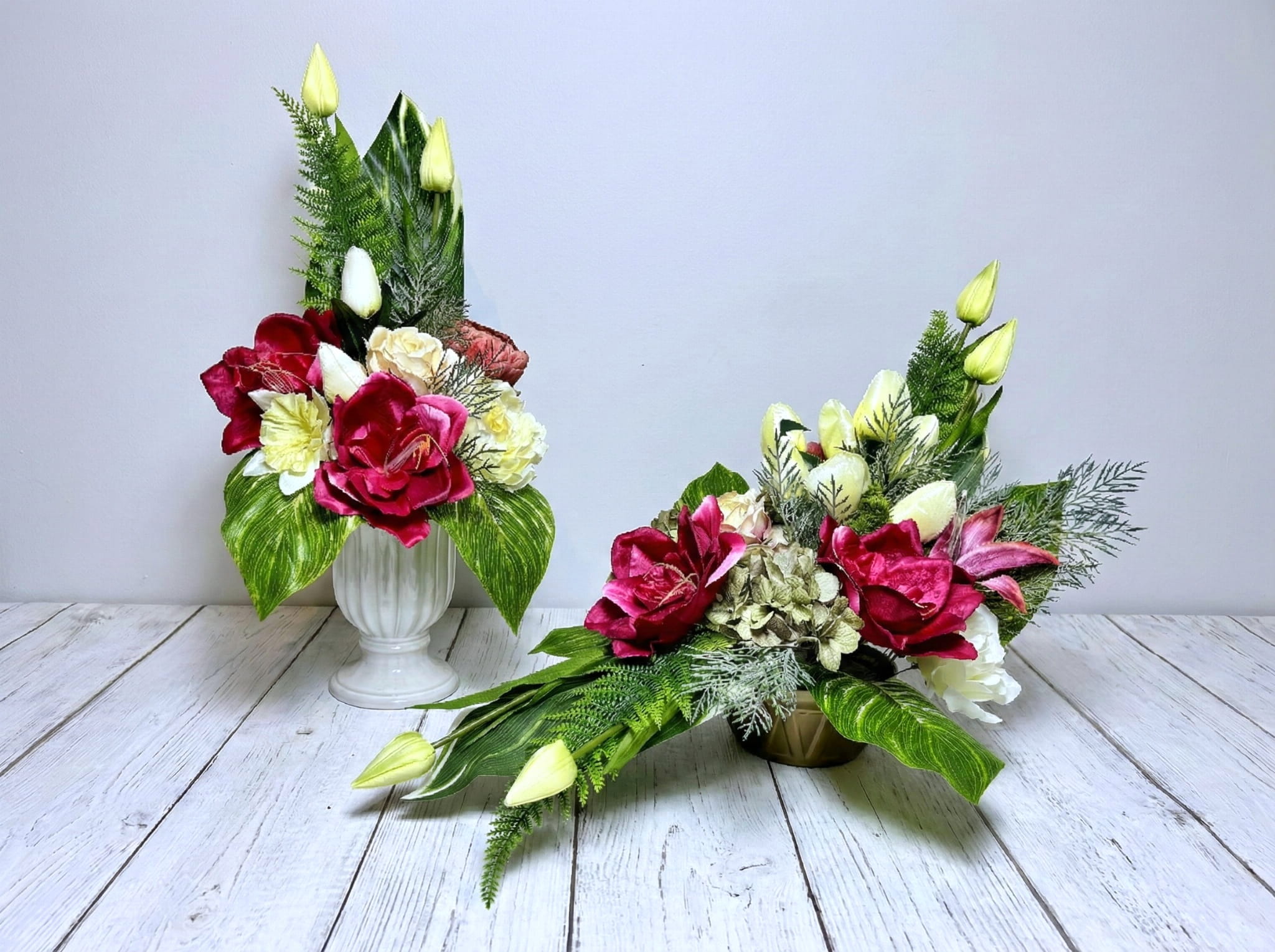 Zdjęcie przedstawia zestaw dwóch dekoracji nagrobnych: podłużny stroik w złotej donicy oraz wysoki bukiet w białym pucharze. Kwiaty są w kolorze kremowym i intensywnej fuksji, głównie piwonie i róże. W kompozycji widoczne są także jasne pąki tulipanów oraz bogata zieleń liści paproci. Produkty ustawione są na białej drewnianej podłodze na tle jasnoszarej ściany.