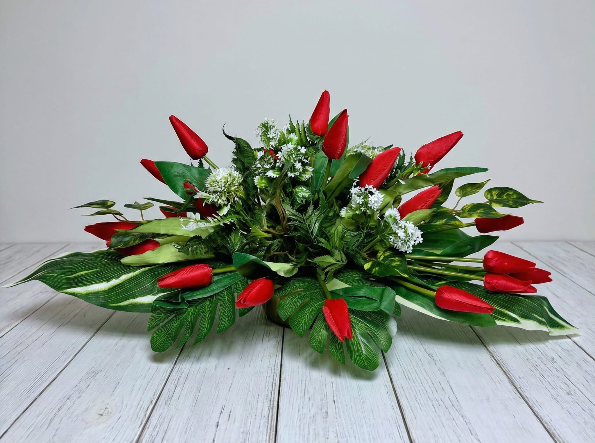 Zdjęcie przedstawia szeroką, niską kompozycję ze sztucznych czerwonych tulipanów w okrągłej złotej donicy. Kwiaty są skierowane w różnych kierunkach i otoczone gęstymi, zielonymi liśćmi o zróżnicowanych kształtach, w tym liśćmi paproci i monstery. Dekoracja ma rozłożysty, horyzontalny kształt o długości 70 cm.