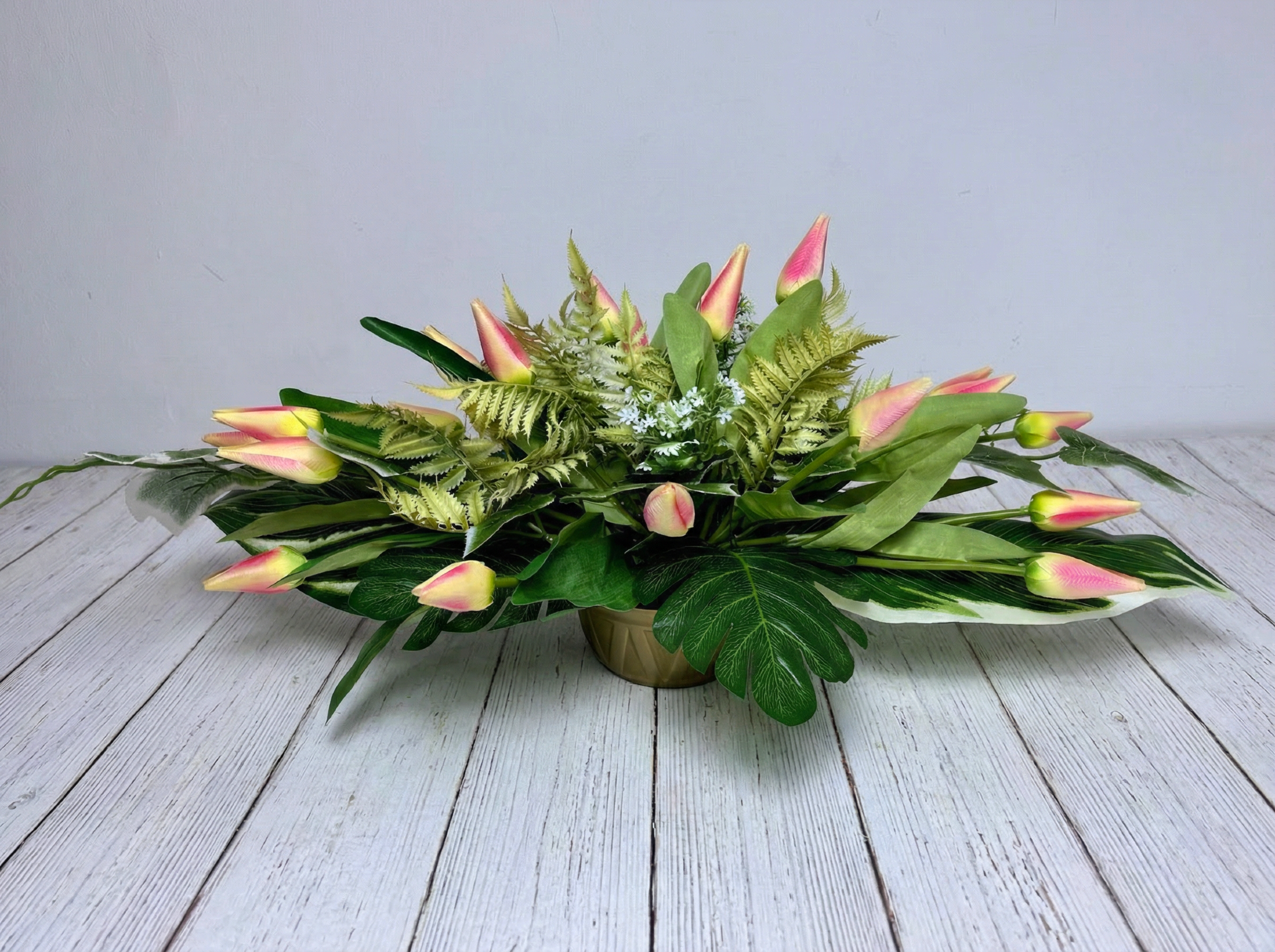 Zdjęcie przedstawia dużą, niską kompozycję ze sztucznych tulipanów o kremowo-różowych płatkach. Kwiaty są gęsto ułożone w okrągłej, złotej donicy i otoczone różnorodnymi zielonymi liśćmi, w tym liśćmi paproci i monstery. Stroik ma podłużny, symetryczny kształt o długości 70 cm. Produkt jest wyeksponowany na białym podłożu, co podkreśla jego pastelową kolorystykę i złote wykończenie naczynia.