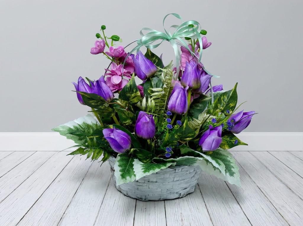 Zdjęcie przedstawia okrągły, bielony kosz wiklinowy wypełniony gęstą kompozycją kwiatową. W środku znajdują się fioletowe tulipany oraz wysokie fioletowe i białe ostróżki. Kwiaty otoczone są bogatą, soczystą zielenią liści i gałązek bukszpanu. Kompozycja o wymiarach 35 cm wysokości i średnicy stoi na jasnym podłożu, prezentując się elegancko i świeżo.