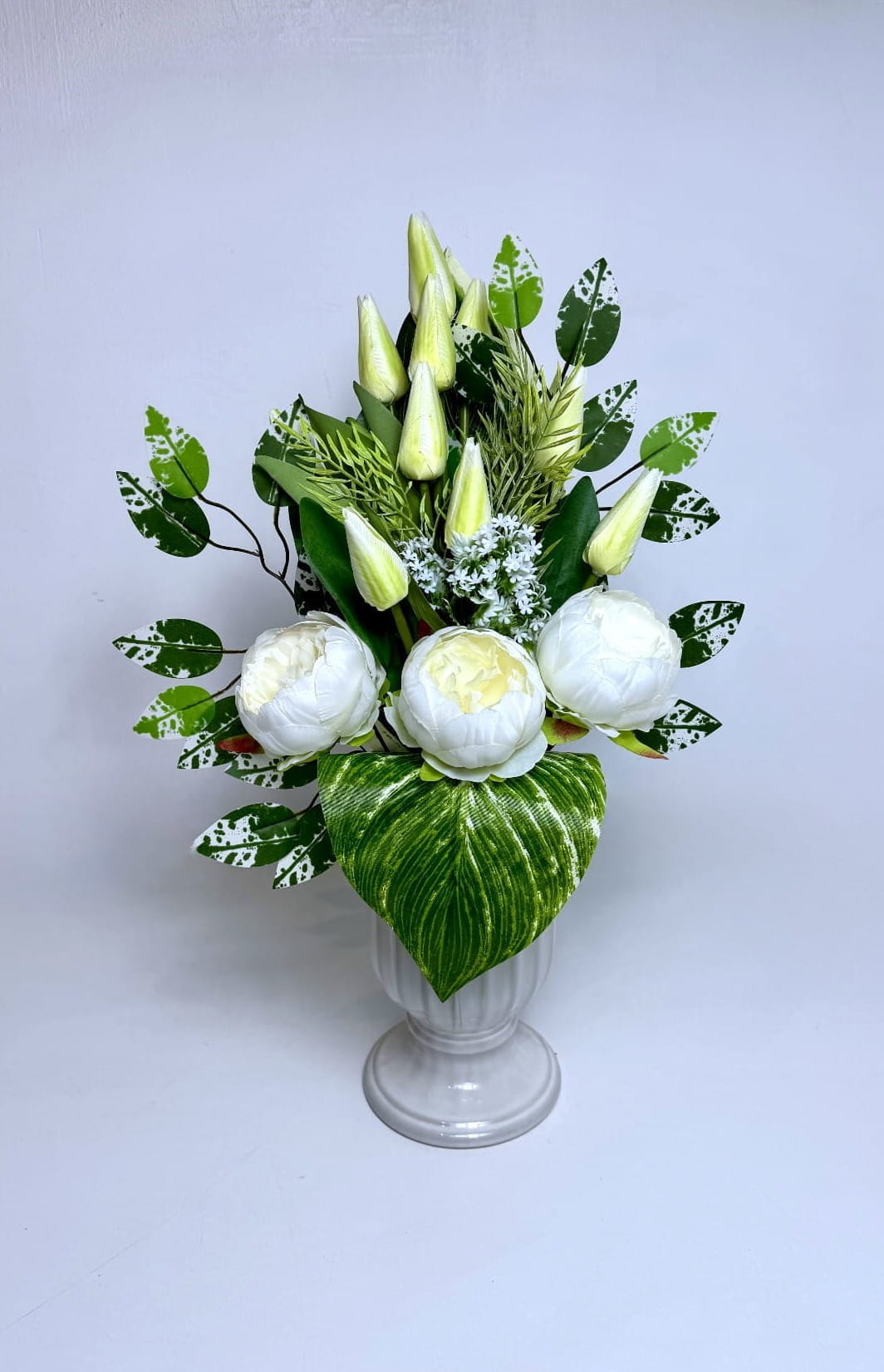 Zdjęcie przedstawia wysoki bukiet nagrobny umieszczony w jasnoszarym, dekoracyjnym wazonie. Dolną część kompozycji tworzą trzy duże, pełne piwonie w kolorze białym z kremowym środkiem. Nad nimi góruje kilkanaście zamkniętych pąków tulipanów w odcieniu jasnej, limonkowej zieleni. Bukiet jest bogato zdobiony liśćmi o charakterystycznym, biało-zielonym nakrapianiu oraz dużym, pasiastym liściem u podstawy. Całość uzupełniają drobne białe kwiatuszki i zielone gałązki dekoracyjne, tworząc smukłą, pionową formę.