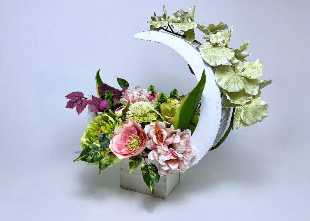 Zdjęcie przedstawia dużą kompozycję kwiatową w białej osłonce w kształcie półksiężyca. Wewnątrz znajdują się gęsto ułożone różowe piwonie, biało-zielone anemony oraz jaskry. Z boku wyrasta długi pęd sylikonowego storczyka w kolorze jasnozielonym. Całość uzupełniają ciemne i zielone liście dekoracyjne. Stroik ma 48 cm wysokości i stoi na białym tle. Produkt wygląda bardzo bogato i elegancko.