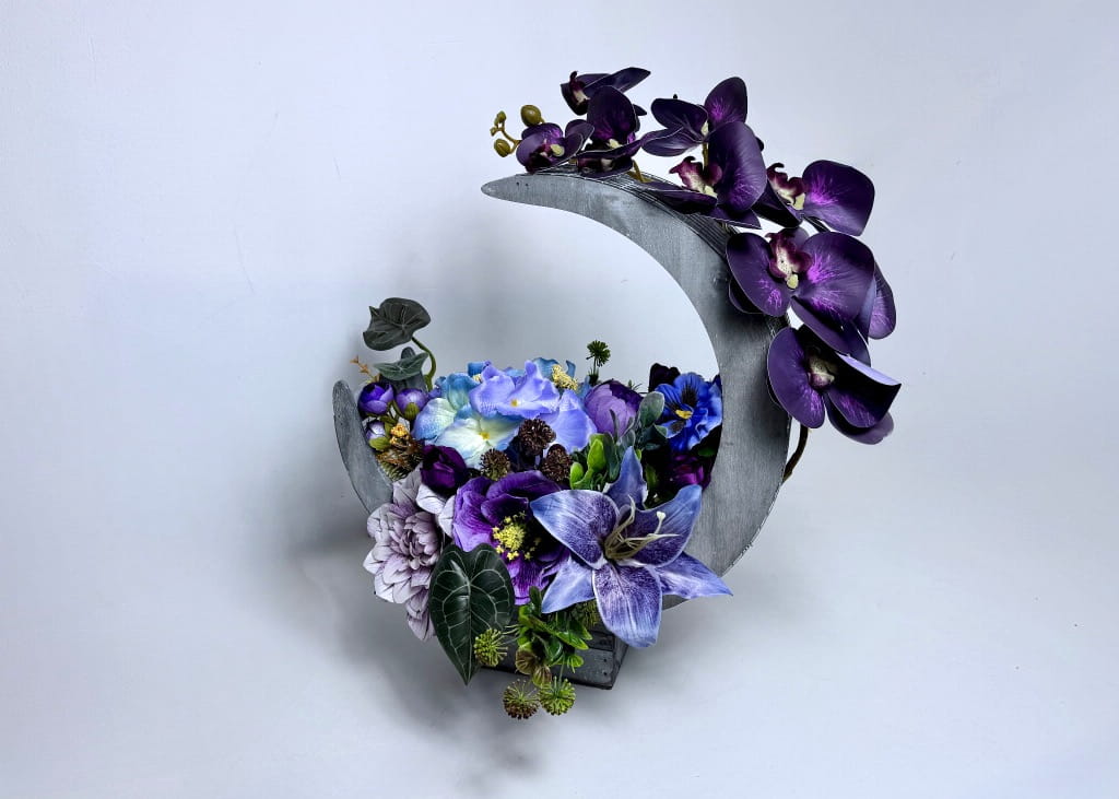 Zdjęcie przedstawia dużą kompozycję kwiatową w czarnym koszu o kształcie półksiężyca. Aranżacja utrzymana jest w tonacji fioletowej i purpurowej, z wykorzystaniem gęstych piwonii, anemonów i jaskrów. Z boku wystaje elegancki, sylikonowy pęd storczyka w kolorze fioletowym. Całość uzupełniają ciemnozielone liście dekoracyjne. Stroik ma 48 cm wysokości i 40 cm długości, stoi na jasnym, jednolitym tle. Produkt wygląda bardzo okazale i luksusowo.