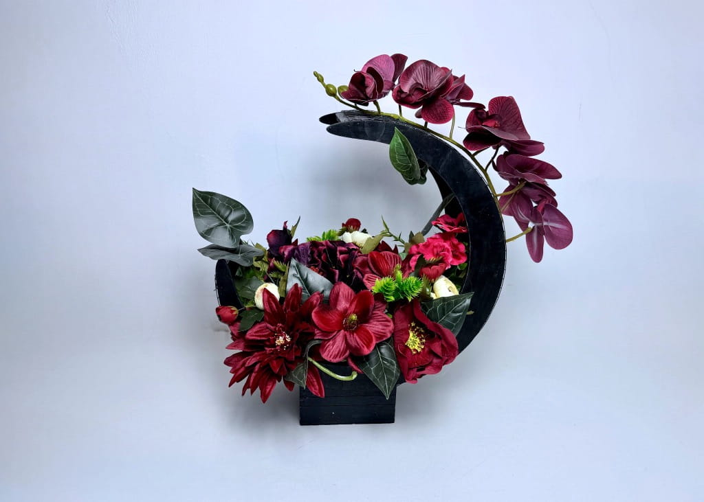 Duża, bogata kompozycja kwiatowa w czarnym, ażurowym koszu w kształcie półksiężyca. Dominują kwiaty w odcieniu ciemnego burgundu i czerwieni: storczyki, dalie i jaskry. Kwiaty są gęsto ułożone, tworząc kaskadową formę. Stroik o wysokości 48 cm, dociążony betonem, umieszczony na jasnym tle. Całość wygląda bardzo ekskluzywnie i solidnie.