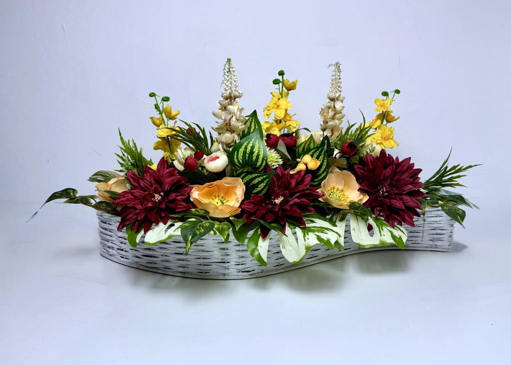 Długa  kompozycja kwiatowa w białym koszu wiklinowym o falistym kształcie. Dominują duże, ciemnoczerwone dalie, strzeliste żółte kwiaty oraz brzoskwiniowe jaskry. Kwiaty otoczone są gęstymi liśćmi monstery z białymi przebarwieniami oraz zielonymi gałązkami. Całość ma 80 cm długości i jest dociążona betonem dla pełnej stabilności.