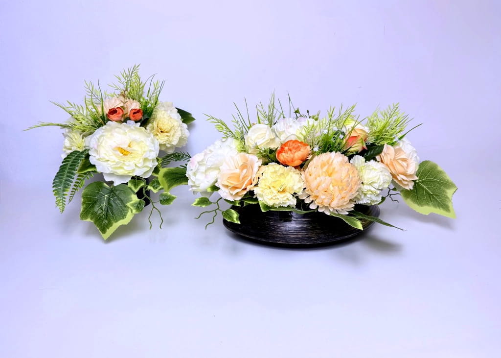 Zestaw kwiatowy w kolorach kremu i brzoskwini w czarnej, owalnej donicy ze złotymi przetarciami. Kompozycja zawiera duże sztuczne piwonie, róże i goździki otoczone gęstymi liśćmi paproci i bluszczu. Obok znajduje się pasujący bukiet do wazonu. Całość ma 70 cm długości i jest dociążona betonem. Zdjęcie na jasnym, neutralnym tle.