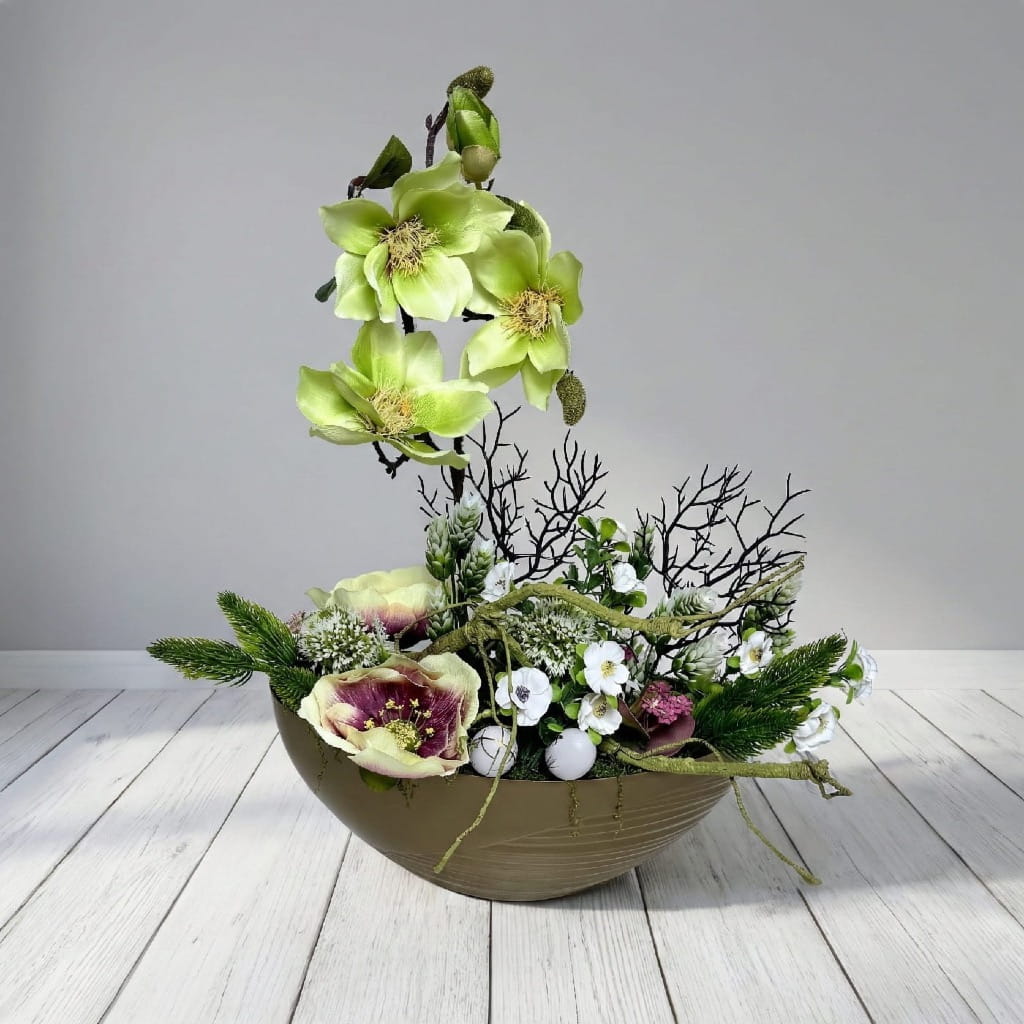 Pionowa kompozycja ze sztucznych kwiatów magnolii i róż w złotej osłonce o kształcie łezki. Kwiaty są w kolorze kremowym i białym, otoczone gęstymi zielonymi liśćmi. Dekoracja ma około 48 cm wysokości i jest osadzona stabilnie w nowoczesnym, połyskującym naczyniu. Produkt znajduje się na tle bielonej drewnianej podłogi i jasnoszarej ściany.