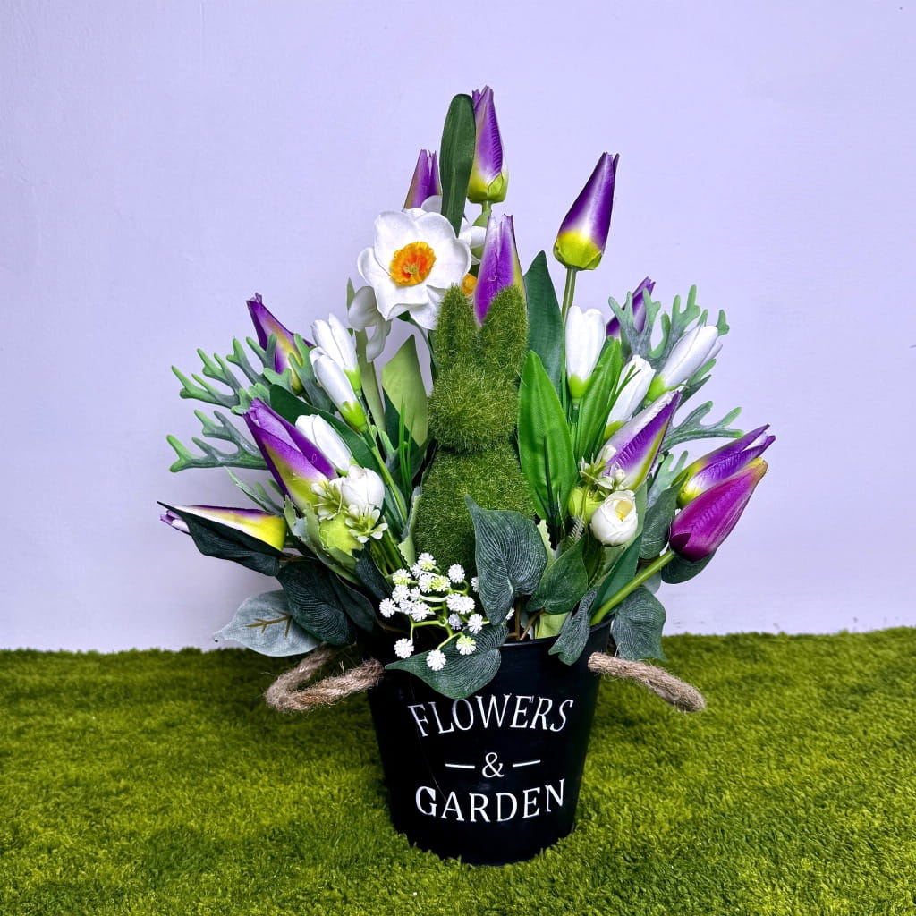 Wiosenna kompozycja w czarnym metalowym kubełku z napisem „FLOWERS &amp; GARDEN”. W centralnej części widać zielonego, mchowego zajączka siedzącego wśród kwiatów. W aranżacji dominują fioletowe tulipany oraz białe narcyzy z pomarańczowymi środkami. Całość dopełniają zielone liście i drobne białe kwiatuszki. Dekoracja ma 40 cm wysokości i jest przedstawiona na tle zielonej, miękkiej trawy i jasnej ściany.