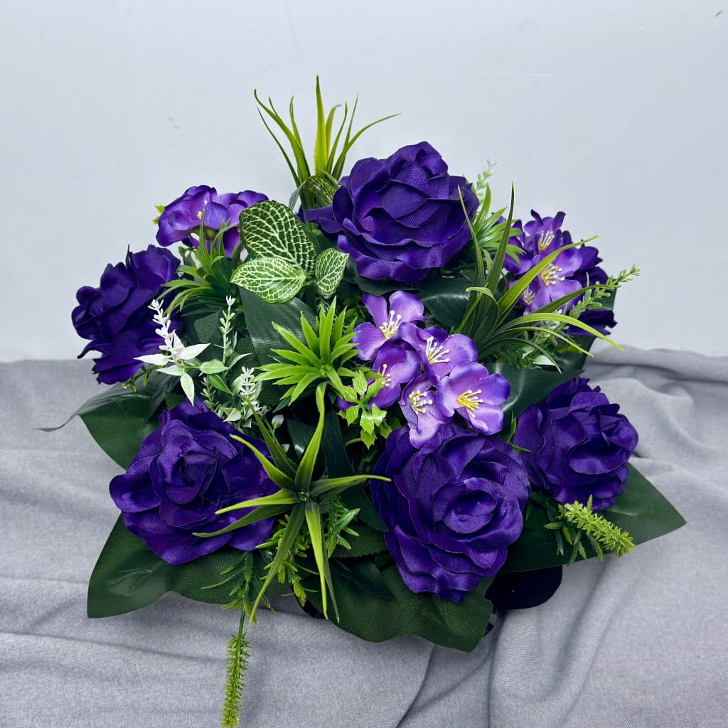 Zdjęcie przedstawia elegancki stroik nagrobny w złotej misie. Głównym punktem kompozycji są duże sztuczne róże w głębokim, fioletowym kolorze o satynowym połysku. Kwiaty są uzupełnione mniejszymi kwiatuszkami w tym samym odcieniu oraz bogatymi dodatkami zielonymi: liśćmi o jasnym unerwieniu, długimi trawami i drobnymi gałązkami dekoracyjnymi. Podstawę stroika zdobi czarna, błyszcząca folia, która podkreśla intensywność fioletu.