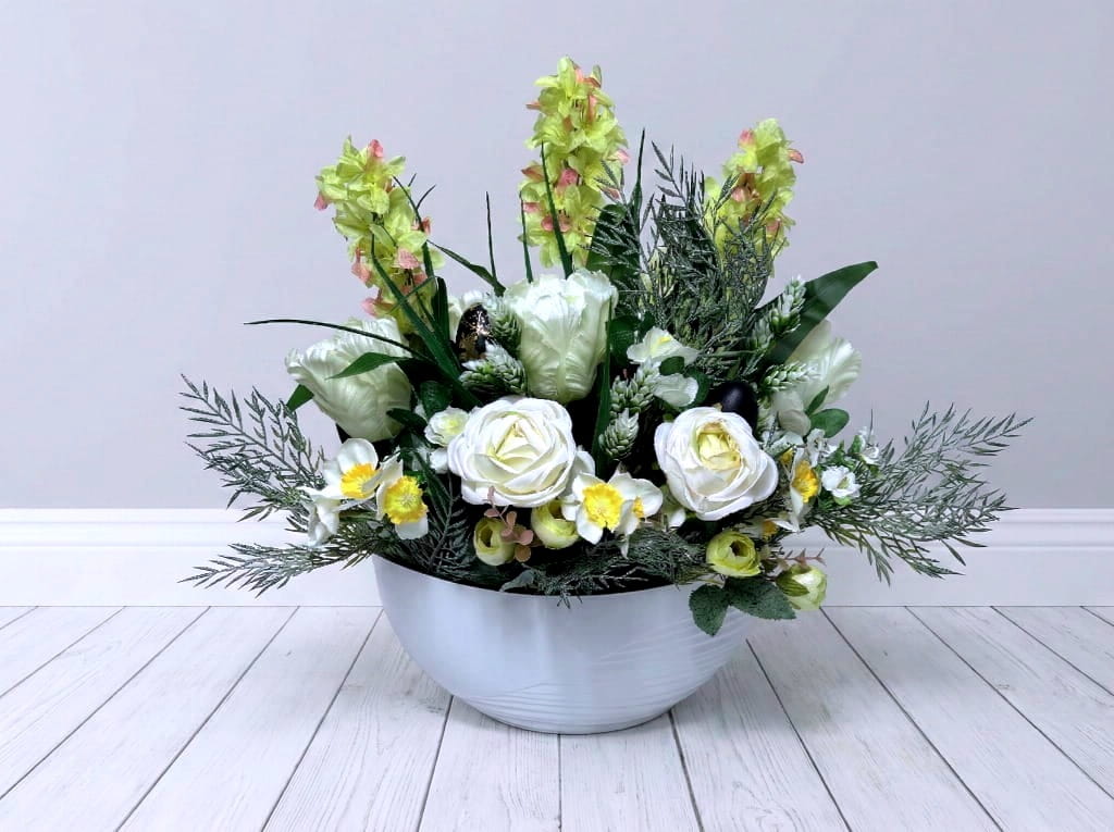 Wysoka, nowoczesna kompozycja kwiatowa w białej donicy o kształcie łezki. Wewnątrz znajdują się białe tulipany, pełne róże i strzeliste hiacynty w tym samym kolorze. Kwiaty uzupełnione są naturalnie zielonymi liśćmi i eukaliptusem. Całość ma smukły, elegancki pokrój i wysokość 45 cm. Kompozycja stoi na jasnej powierzchni, prezentując czysty i luksusowy charakter.