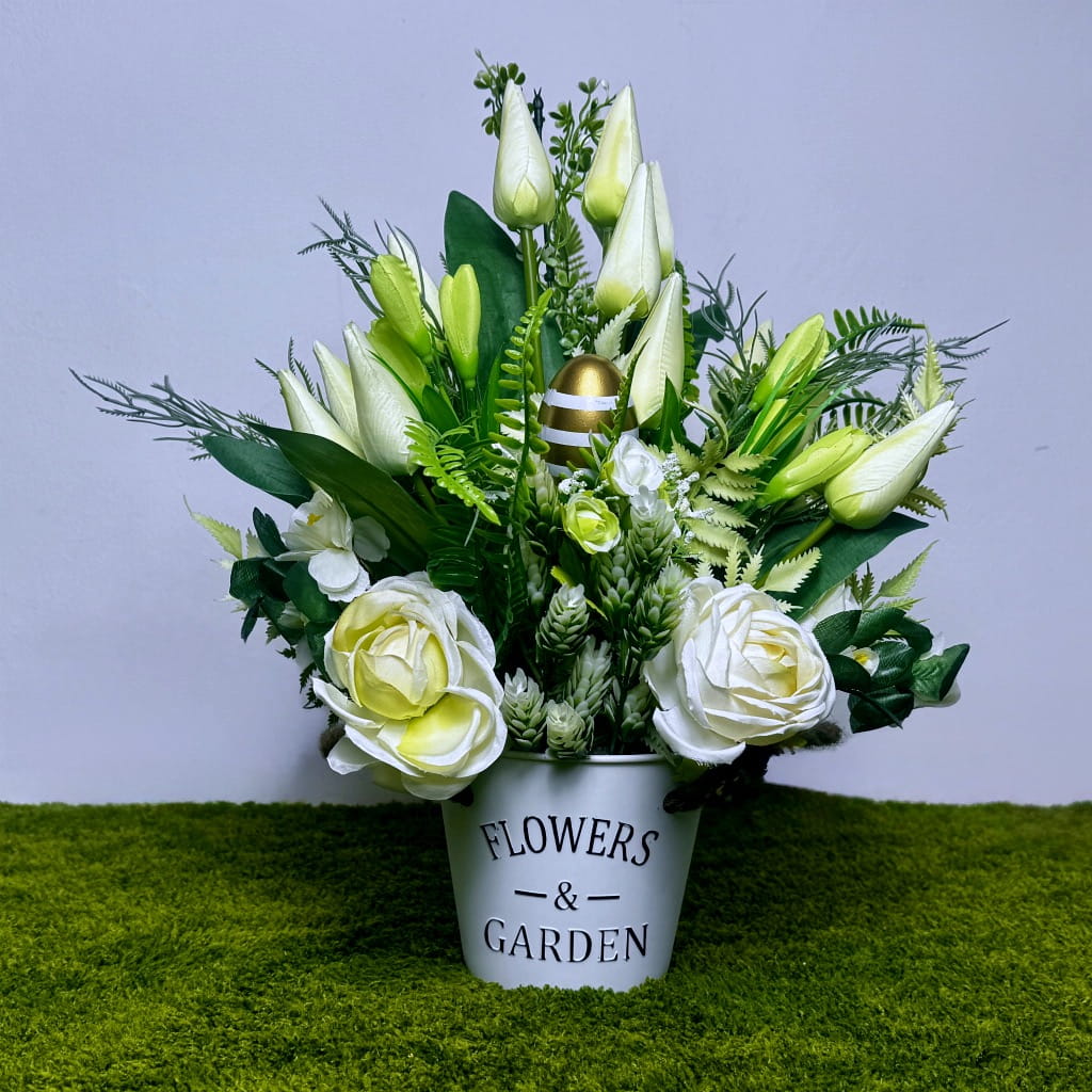 Zdjęcie przedstawia pionową, wiosenną dekorację w białej plastikowej osłonce z napisem "Flowers &amp; Garden". Wewnątrz gęste białe tulipany, róże i jaskry otoczone liśćmi monstery oraz paproci. W centrum znajduje się duże złote jajko z białymi pasami. Kompozycja ma około 40 cm wysokości, jest dociążona i stabilna. Całość stoi na zielonej trawie na jasnym tle. Kolory są czyste, słoneczne i bardzo eleganckie, idealne do domu lub jako trwała ozdoba grobu.