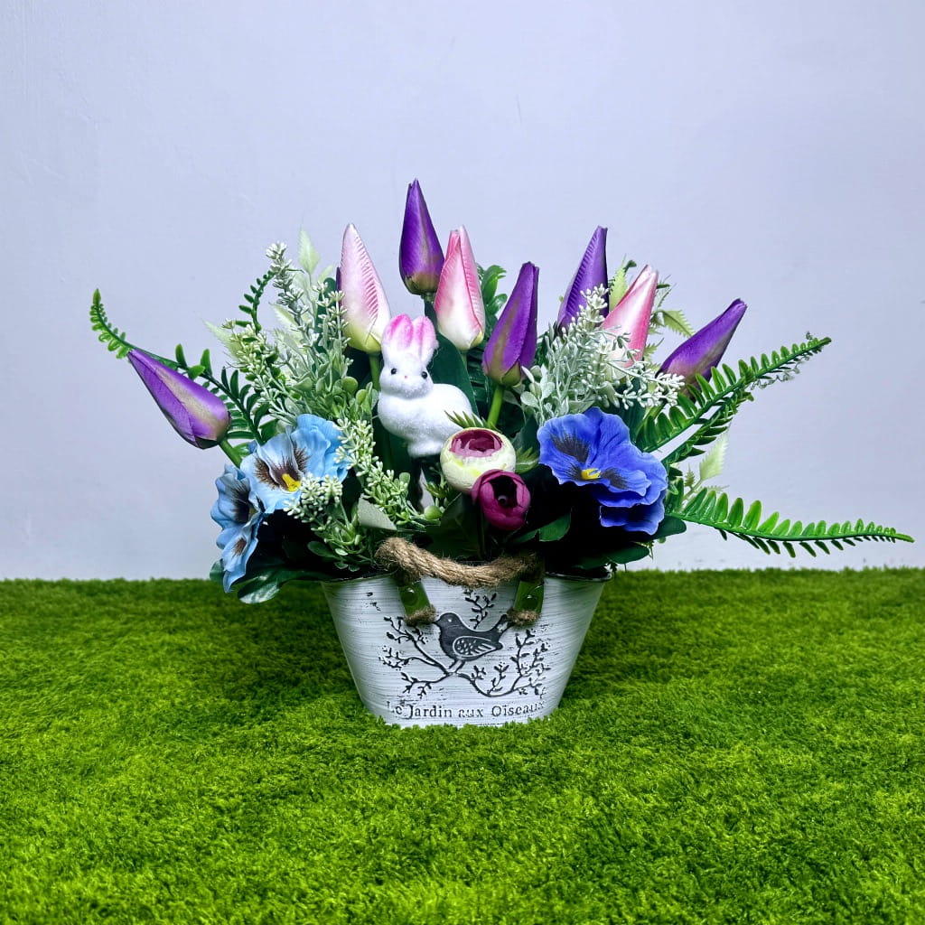 Zdjęcie przedstawia wiosenną kompozycję kwiatową w białej, metalowej osłonce z motywem ptaka i sznurkowymi uchwytami. Wewnątrz gęsto ułożone fioletowe tulipany, niebieskie bratki oraz drobne fioletowe kwiaty akcentowe. W środku dekoracji siedzi mała figurka białego, puszystego zająca. Kwiaty otoczone są zielonymi liśćmi paproci i gałązkami eukaliptusa. Wysokość stroika wynosi około 30 cm. Całość zaprezentowana na zielonym podłożu imitującym trawę na jasnym tle. Kolory są żywe i kontrastowe.