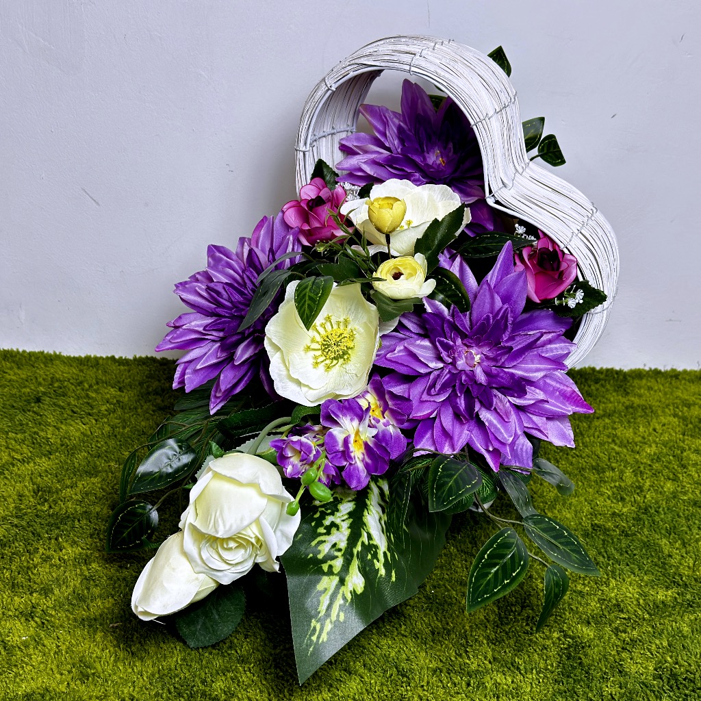 Zdjęcie przedstawia asymetryczną kompozycję kwiatową osadzoną w białym, plecionym koszu w kształcie serca, leżącym na boku. Wewnątrz znajdują się okazałe fioletowe dalie, kremowo-białe anemony z żółtymi środkami, kremowe i fuksjowe róże, fioletowe ostróżki oraz kremowe jaskry. Całość otaczają gęste zielone liście paproci.
