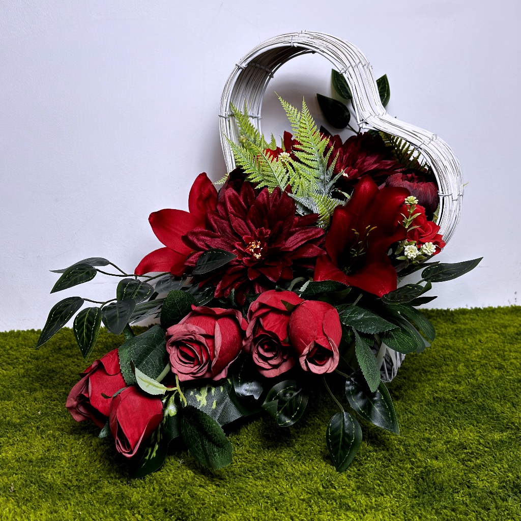 Zdjęcie przedstawia poziomą kompozycję w białym koszu w kształcie serca. Dominują intensywnie czerwone lilie, dalie i róże. Całość uzupełniają zielone liście paproci oraz gałązki ozdobne. Kompozycja jest gęsta i ułożona asymetrycznie wewnątrz plecionego naczynia, które stoi na zielonym podłożu imitującym trawę.