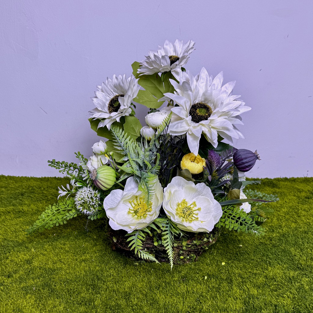 Zdjęcie przedstawia pionową kompozycję kwiatową w owalnym koszyku wykonanym z zielonego mchu. Wewnątrz znajdują się białe słoneczniki, białe anemony, drobne jaskry, strzelisty czosnek ozdobny oraz fioletowe i zielone główki maków (makówki). Całość otaczają gęste zielone liście paproci i eukaliptusa. Stroik ma jasną, pastelową kolorystykę i naturalny wygląd. Wysokość kompozycji to 40 cm. Podstawa jest stabilna, a kwiaty ułożone gęsto.