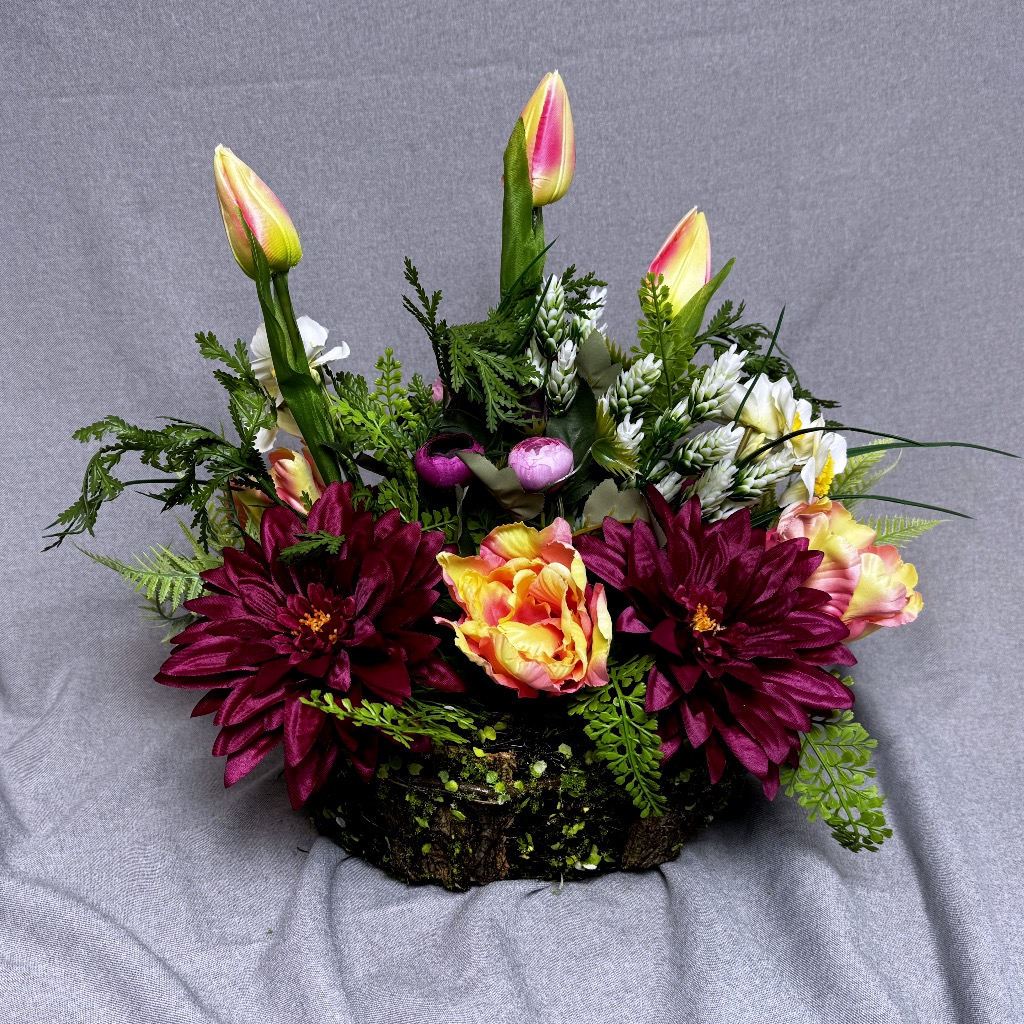 Zdjęcie przedstawia okrągłą, gęstą kompozycję kwiatową w zielonym koszyczku wykonanym z mchu. Wewnątrz znajdują się różowe dalie, kremowo-różowe róże, jaskry oraz tulipany. Całość uzupełniają liczne liście paproci i gałązki eukaliptusa. Stroik jest dwustronny, ma pastelową kolorystykę i naturalny wygląd. Wysokość kompozycji to ok. 40 cm. Podstawa jest stabilna i dociążona.