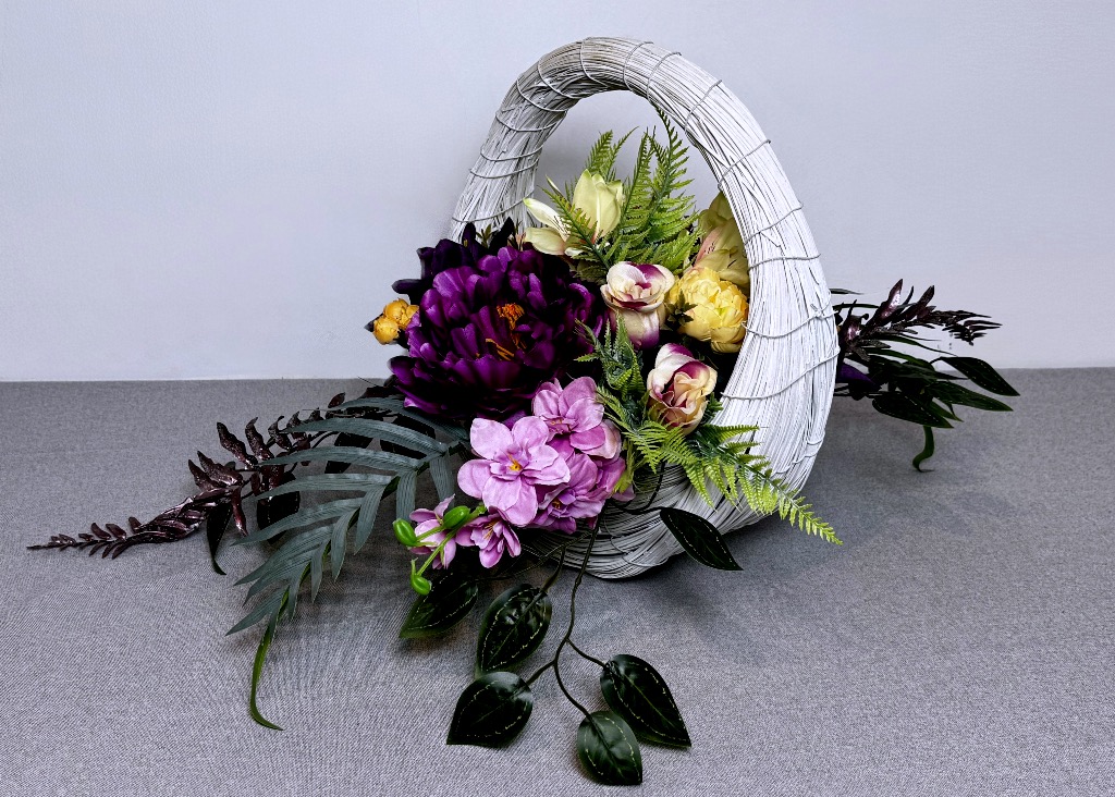 Kolorowa kompozycja kwiatowa w okrągłym wiklinowym koszu z pałąkiem. Wewnątrz znajdują się purpurowe i żółte piwonie, strzeliste różowe ostróżki, ciemnofioletowe lilie oraz kremowe pąki róż. Bogata zieleń z liśćmi paproci i eukaliptusa wypełnia boki. Podstawa z naturalnej, jasnej wikliny stoi stabilnie. Całość ma radosny, wielobarwny charakter i gęstą strukturę.