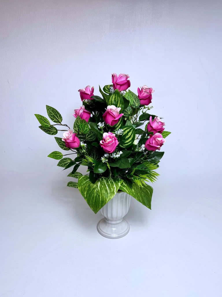 Stożkowy bukiet nagrobny do wazonu. Składa się z pąków róż o intensywnie różowych płatkach zewnętrznych i jasnokremowych środkach. Kwiaty otoczone są gęstymi liśćmi: ciemnozielonymi liśćmi róży oraz jasnymi listkami z żółtym unerwieniem. U podstawy znajdują się dwa duże, szerokie liście z wyraźnymi białymi pasami. Między różami widać drobne białe akcenty kwiatowe. Całość osadzona w czarnym wkładzie. Wymiary: wysokość 50 cm, szerokość 35 cm.