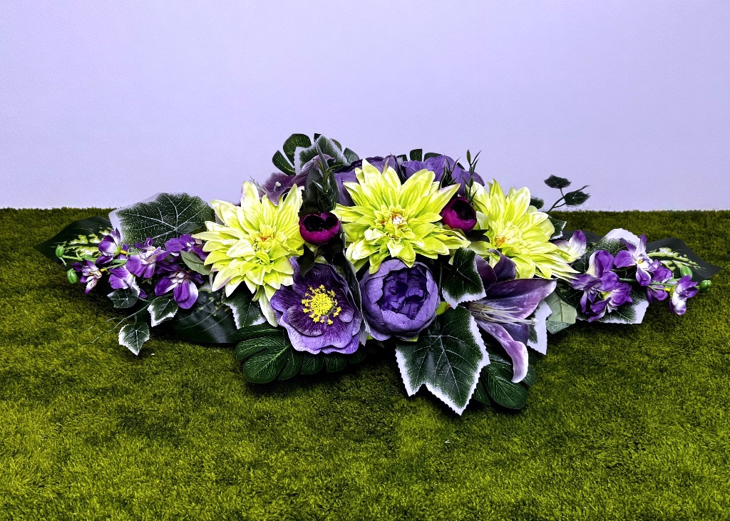 Podłużna wiązanka na grób w złotej donicy. W centralnej części znajdują się trzy duże seledynowe dalie, otoczone fioletowymi piwoniami i liliami. Na boki wychodzą fioletowe ostróżki. Liście są zielone z białymi brzegami, gęsto wypełniające spód. Kompozycja ma 80 cm długości i leży stabilnie na zielonym podłożu. Kolory są żywe i kontrastowe.