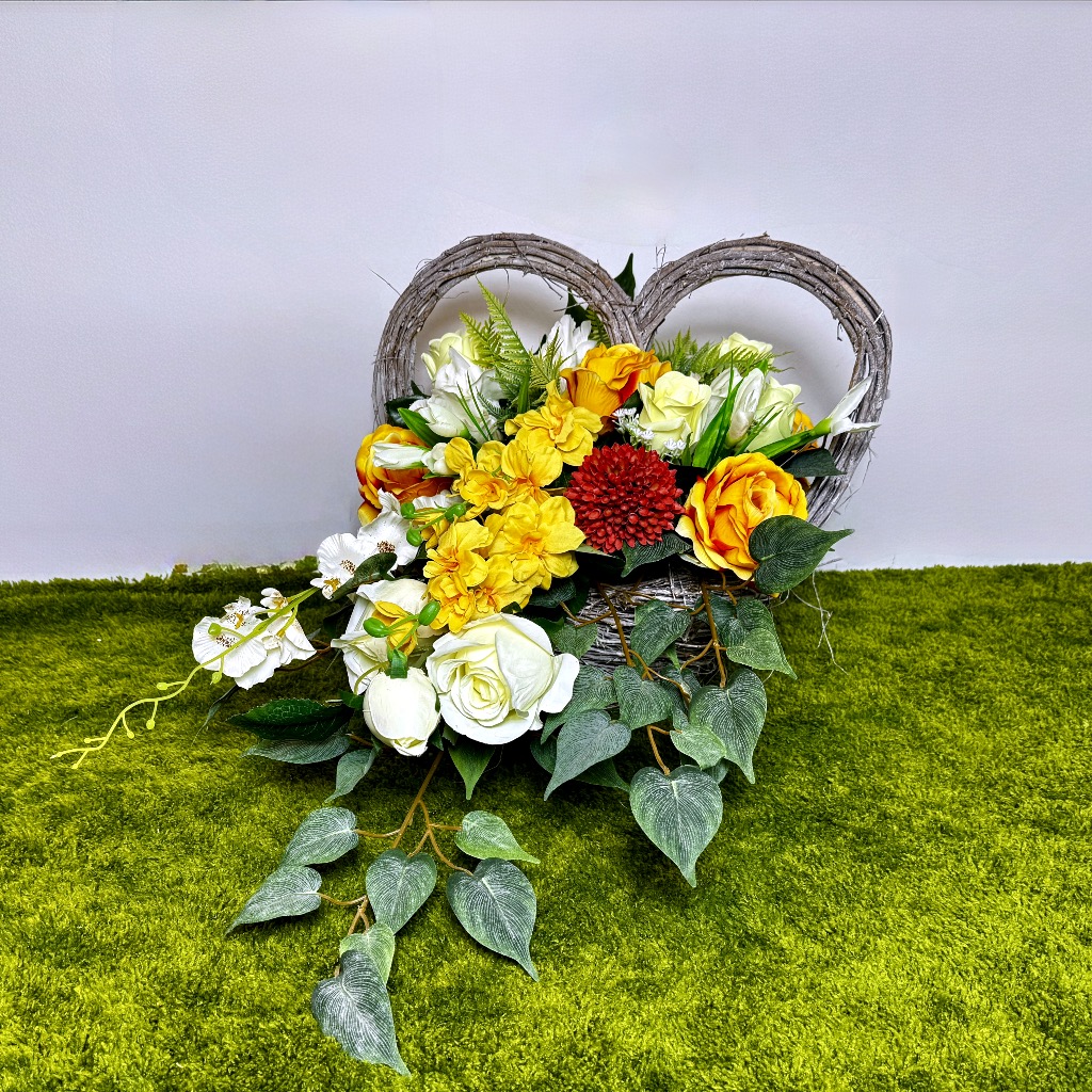 Duży, biały wiklinowy kosz w kształcie serca, wypełniony gęstą kompozycją kwiatową. Dominują jasne, żółte róże i białe tulipany. Charakterystycznym elementem są czerwone kule czosnku ozdobnego oraz wysokie ostróżki. Całość otaczają zielone liście paproci. Kompozycja jest długa, niska i leżąca. Podstawa jest stabilna.