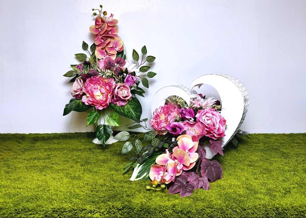 Okazały zestaw różowych kwiatów na cmentarz. Składa się z białego kosza w kształcie serca wypełnionego gęsto różami i storczykami oraz pasującego bukietu w wazonie. Kwiaty mają różne odcienie różu i wyglądają jak prawdziwe. Konstrukcja serca jest dwustronna i bardzo długa, mierzy 90 cm. Całość stoi na zielonej trawie, prezentując się bardzo elegancko i solidnie.