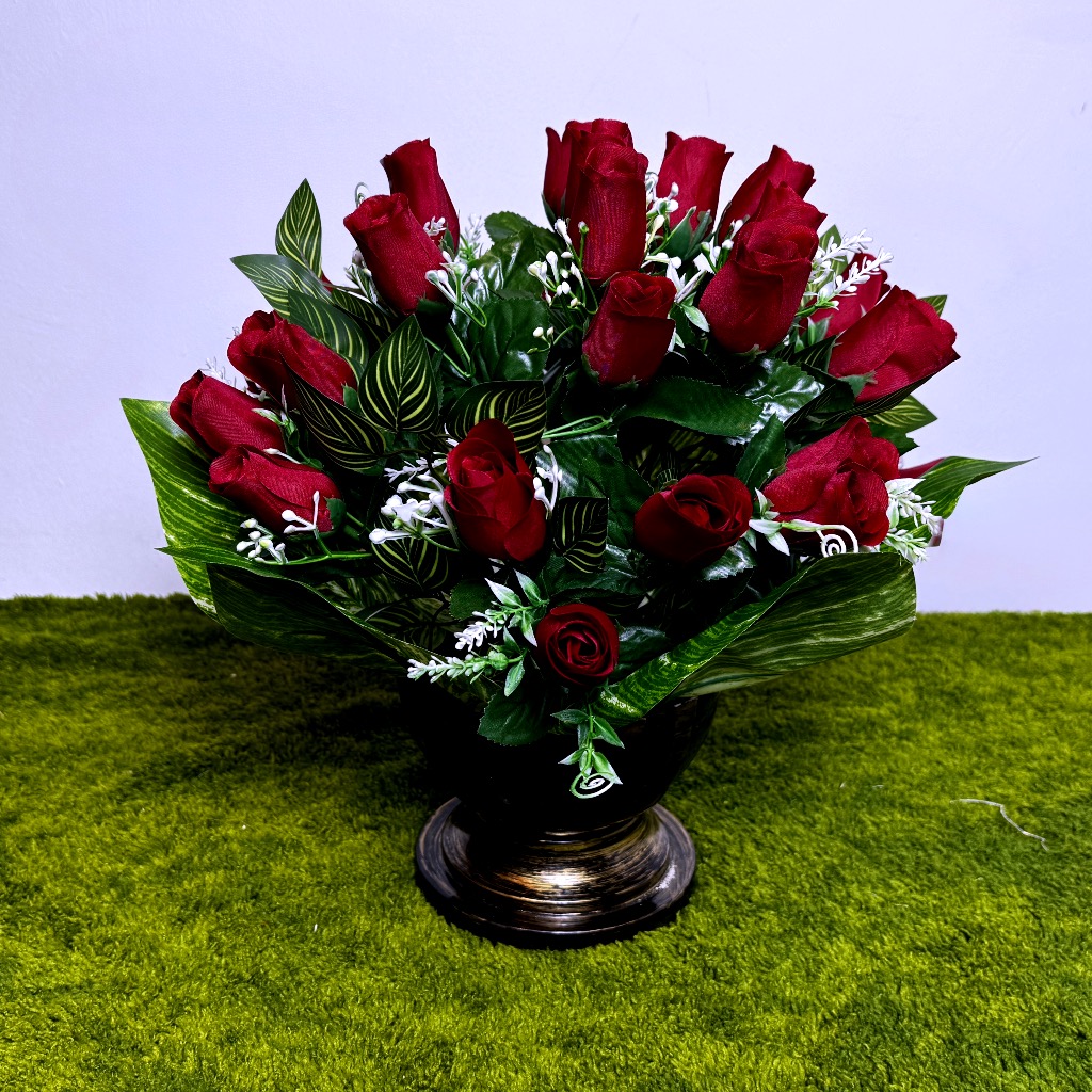 Gęsty bukiet czerwonych róż w czarnym naczyniu typu puchar ze złotymi wzorami. Donica ma stabilną nogę i jest dociążona betonem. Róże otaczają zielone liście oraz długie pnącza bluszczu z jasnymi brzegami, które spływają na bok. Kompozycja jest szeroka, ma 40 cm wysokości. Kwiaty mają realistyczną fakturę i głęboki, czerwony kolor. Produkt stoi na zielonej trawie.