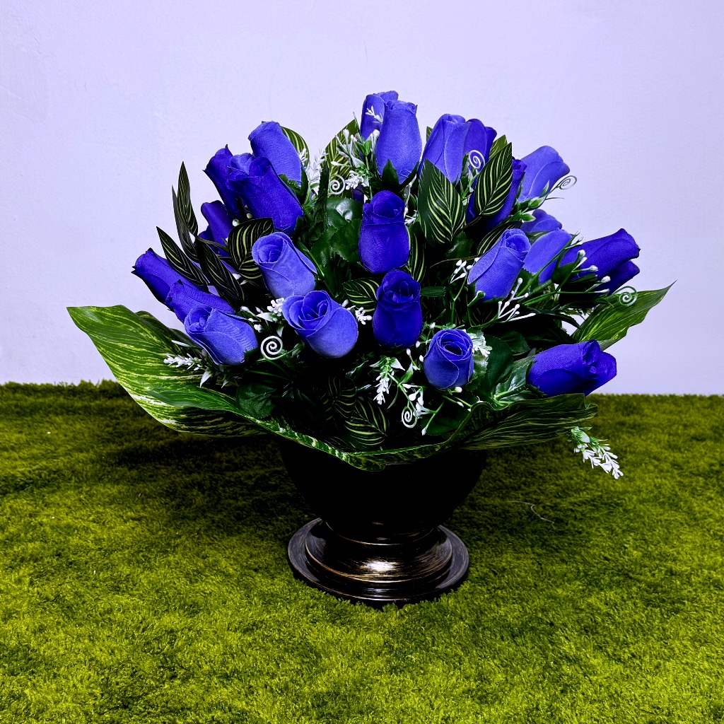 Okrągły bukiet róż w kolorach ciemnoniebieskim i błękitnym. Kwiaty są gęsto ułożone w czarnym naczyniu na nodze ze złotymi wzorami. Pomiędzy różami widać ciemnozielone liście i małe białe kropki kwiatuszków. Kompozycja ma wysokość 40 cm i jest dociążona masą betonową. Całość wygląda realistycznie i solidnie, stoi na zielonym podłożu imitującym trawę.
