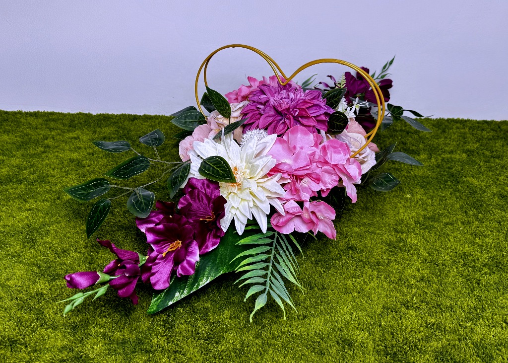 Dwustronna, długa wiązanka nagrobna umieszczona w złotym metalowym sercu. Kompozycja składa się z malinowych i kremowych dalii, różowych mieczyków, anemonów oraz róż. Kwiaty są gęsto ułożone, otoczone dużymi zielonymi liśćmi monstery i bluszczu. Całość ma 80 cm długości, jest dociążona i stabilna. Kolory są żywe: od intensywnej maliny po delikatny krem i róż.